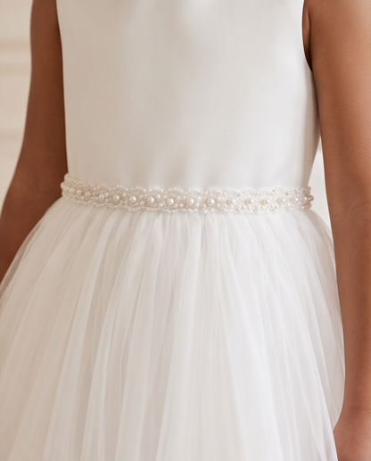 Close-up of a child wearing a white sleeveless dress with a fitted bodice, a pearl and bead waistband, and a layered tulle skirt.