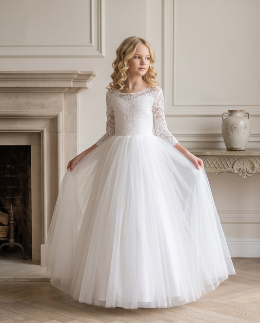 A young girl wearing a white ball gown wedding dress with lace bodice and a full tulle skirt, standing in a classical room with a fireplace and holding the sides of her dress outwards.