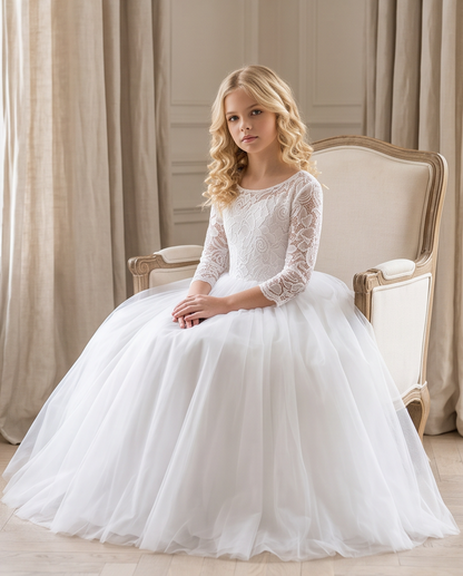 A young girl with blonde wavy hair sits in an elegant cream-colored armchair wearing a white ball gown with a lace bodice and a full tulle skirt, set against a soft background of beige curtains and paneled walls.