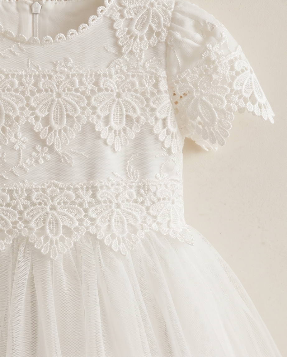 Close-up of a white children's dress with floral lace applique, short puff sleeves, and a tulle skirt against a plain light beige background.