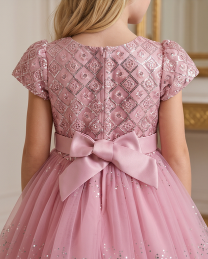 A child wearing a pink party dress, shown from the back. The dress features a sparkling sequined and embroidered bodice with short puff sleeves, a large satin bow at the waist, and a voluminous tulle skirt with subtle silver glitter.