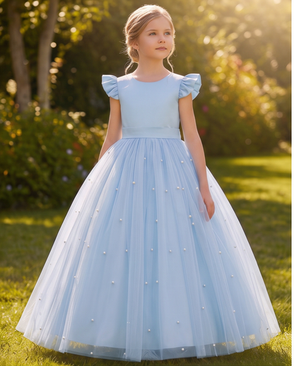 A young girl stands outdoors on grass wearing a light blue ball gown with ruffled cap sleeves, a fitted bodice, and a full tulle skirt decorated with small pearl-like embellishments.