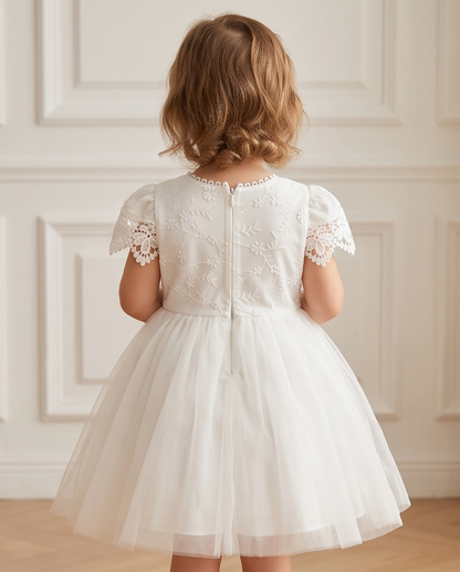 A young girl viewed from behind wearing a white tulle dress with floral lace embroidery on the back and short lace sleeves, standing in front of a cream paneled wall.