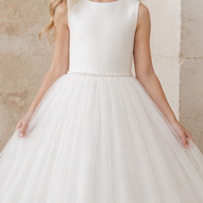 A child wearing a sleeveless white dress with a fitted bodice, a pearl-like waistband, and a full tulle skirt, standing against a softly textured beige wall.