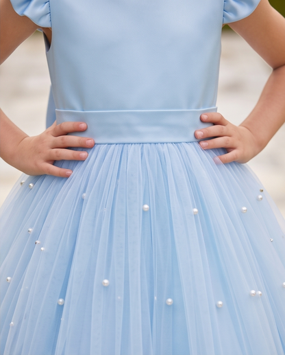 A close-up of a child wearing a light blue dress, showing the waistband, short sleeves, and a full tulle skirt decorated with small pearl-like beads.