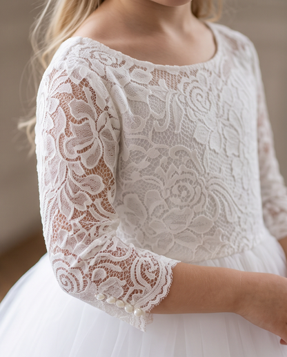 A close-up of a young girl wearing a white lace and tulle dress, showing the detailed floral lace bodice and three-quarter sleeves with pearl accents.