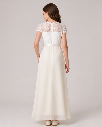 A woman viewed from the back wearing a floor-length ivory dress with a lace lace-embroidered bodice, cap sleeves, and a flowing chiffon skirt, set against a solid light gray studio background.