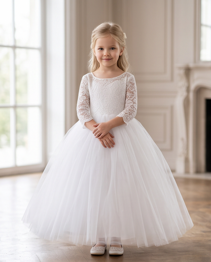 A young girl with blonde hair stands in a bright, elegant room wearing a formal white flower girl dress with a lace bodice, three-quarter lace sleeves, and a full tulle skirt.