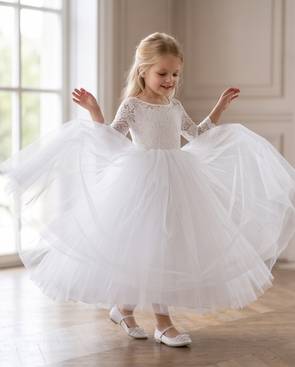 A young girl with blonde hair wearing a white lace long-sleeve first communion dress with a voluminous tulle skirt, twirling on a wooden floor in a bright room.