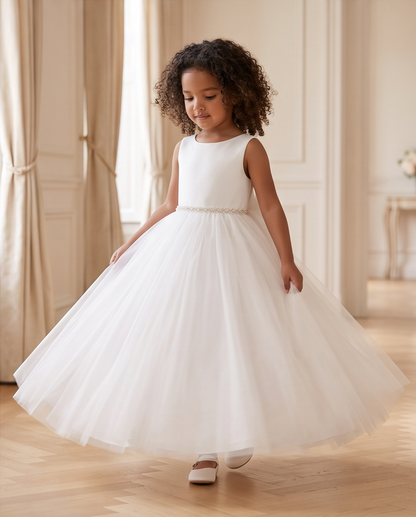 A young girl with curly dark hair wearing a sleeveless white tulle dress with a beaded waistband, standing in a bright, elegant room with beige walls and wood floors.