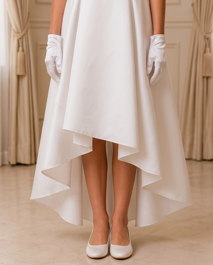 A high-low wedding dress with a flowing white satin skirt, styled with white gloves and white heels, shown on a model in an elegant indoor setting.