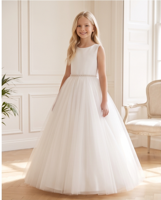 A young girl with long blonde hair stands in a bright, elegant room wearing a sleeveless white formal dress with a fitted bodice, a delicate beaded waistband, and a full tulle skirt.
