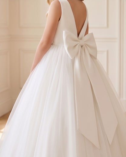 A child wearing a sleeveless white dress from the back, featuring a large satin bow at the back of the V-neckline and a full tulle skirt, set against a soft beige paneled wall.