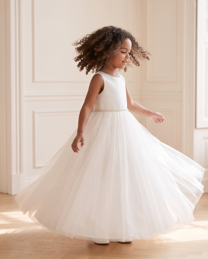 A young girl with curly brown hair wearing a sleeveless white first party dress with a tulle skirt, twirling in a brightly lit room with cream paneled walls and wood floors.